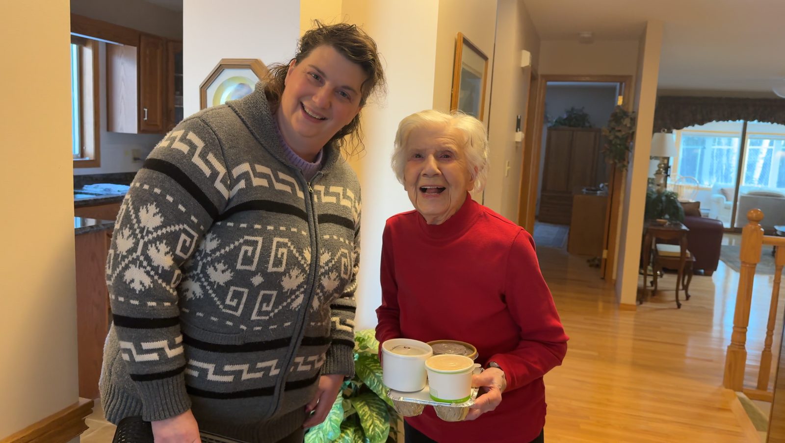 A smiling client at home holding her soup and lunch, beside a volunteer